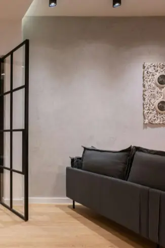 La imagen muestra un espacio interior minimalista y moderno. A la izquierda, destaca una puerta de diseño industrial con marco de metal negro y cristales, que añade un toque contemporáneo y conecta con otra habitación. El fondo es una pared lisa de color gris claro, que contribuye a la sensación de amplitud y simplicidad del espacio.  A la derecha, se encuentra un sofá de líneas rectas en tono gris oscuro, con cojines que proporcionan comodidad y un diseño funcional. El suelo es de madera clara, lo que aporta calidez y contrasta sutilmente con los tonos neutros predominantes en el resto del ambiente. En la pared, se aprecia un elemento decorativo con diseño calado, que añade un toque de detalle y sofisticación al conjunto.  La iluminación proviene de focos empotrados en el techo, que realzan los detalles de los materiales y colores. El espacio combina funcionalidad y diseño, ideal para un ambiente relajado y elegante.