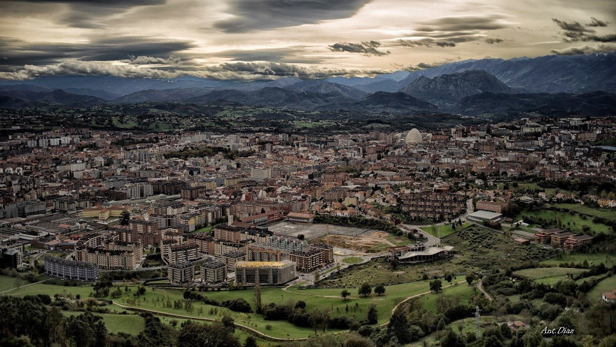 La imagen muestra una vista panorámica de Oviedo, la capital del Principado de Asturias, en un entorno urbano rodeado de paisajes naturales. En primer plano, se pueden apreciar edificios residenciales y zonas verdes que destacan por su integración en el paisaje urbano. Al fondo, las montañas enmarcan la ciudad, creando un contraste entre la arquitectura y la naturaleza, con un cielo parcialmente cubierto que aporta dramatismo a la escena. Es una representación visual de la armonía entre lo urbano y lo natural que caracteriza a esta ciudad del norte de España.