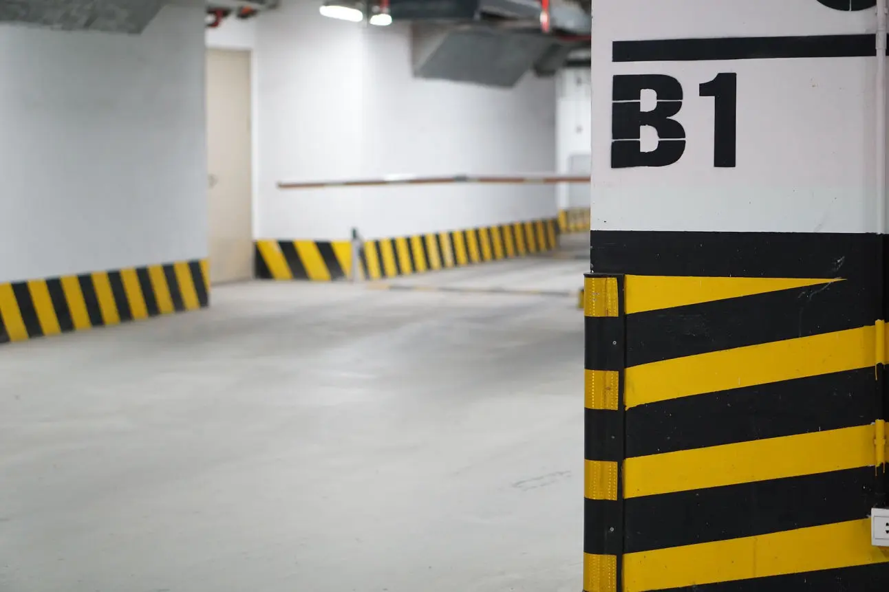 La imagen muestra una zona de aparcamiento subterráneo, destacando un diseño funcional y señalización de seguridad. En primer plano, aparece una columna marcada con franjas diagonales negras y amarillas, típicas para alertar sobre obstáculos y prevenir accidentes. En la parte superior de la columna se encuentra el indicador “B1”, que probablemente señala el nivel o planta del parking.  El fondo presenta un espacio amplio y bien iluminado, con paredes blancas y líneas de señalización en el suelo y las paredes que guían a los conductores. El diseño limpio y ordenado, junto con los elementos de seguridad, transmite un ambiente práctico y estructurado, ideal para el tránsito y estacionamiento de vehículos.