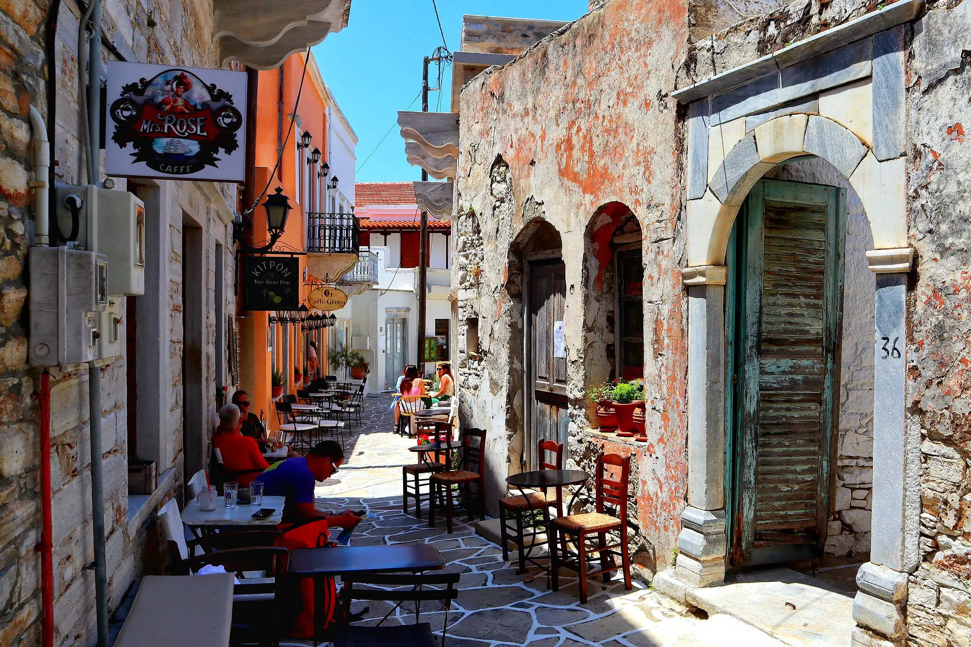 La imagen muestra una calle pintoresca con un encanto rústico y tradicional. A la izquierda, se ven mesas y sillas dispuestas al aire libre, probablemente de un café o restaurante, donde algunas personas disfrutan del ambiente relajado. Las fachadas de los edificios tienen un estilo mediterráneo, con colores cálidos como el naranja y detalles envejecidos que resaltan el carácter histórico del lugar. A la derecha, se aprecian arcos de piedra, puertas de madera desgastada y macetas con flores que aportan un toque de vida y frescura. La calle, con un suelo de piedra cuidadosamente trabajado, invita a recorrerla bajo un cielo despejado, transmitiendo una sensación de tranquilidad y autenticidad.