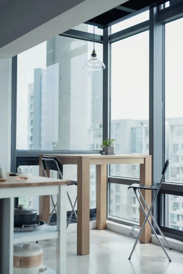 La imagen muestra un rincón de comedor o zona de trabajo con un diseño moderno y minimalista, ubicado en lo que parece ser un apartamento con amplios ventanales de cristal que permiten una vista de la ciudad y proporcionan abundante luz natural. El espacio está decorado con una mesa alta de madera clara y dos sillas plegables metálicas con asiento negro.  En el centro, colgando del techo, hay una lámpara de estilo industrial con pantalla de vidrio transparente, complementando el diseño sencillo del lugar. En una esquina de la mesa, se encuentra una pequeña planta en una maceta que añade un toque de frescura al ambiente.  A la izquierda de la imagen, se distingue parcialmente un carrito o estante blanco con utensilios de cocina, lo que sugiere que este espacio podría estar integrado con una cocina. El suelo es de un tono claro y pulido, acentuando el estilo limpio y contemporáneo del entorno.
