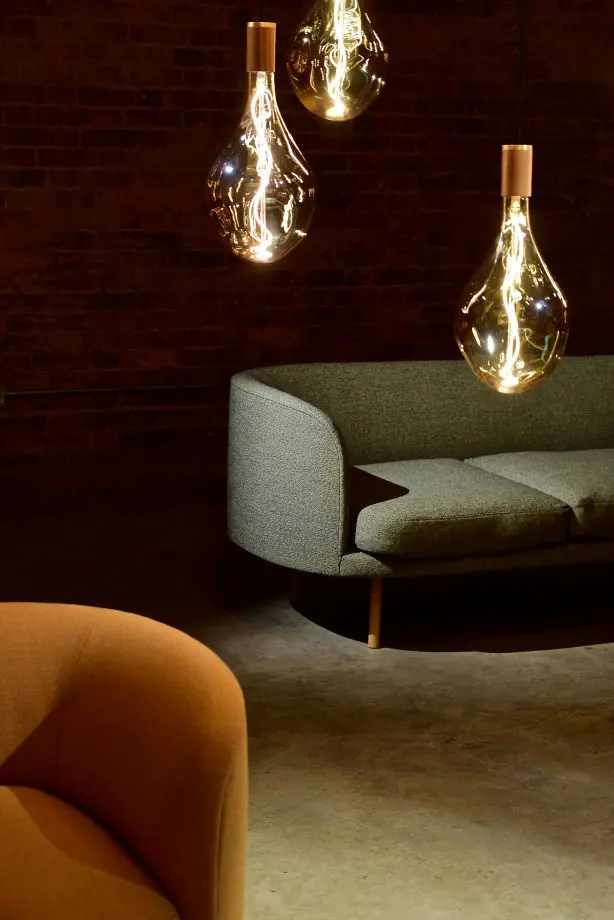 La imagen muestra un ambiente interior acogedor y elegante, con un estilo moderno y cálido. El fondo está compuesto por una pared de ladrillo oscuro que añade textura y contraste al espacio. En el centro, destacan tres lámparas colgantes con bombillas de filamento visible, encerradas en pantallas de vidrio transparente con forma de lágrima y acabados dorados en la parte superior. Estas lámparas emiten una luz cálida que realza la atmósfera íntima del lugar.  En el lado derecho, se observa un sofá de diseño curvo tapizado en tela gris verdosa, con patas de madera que elevan ligeramente el mueble del suelo. En la esquina inferior izquierda, se distingue parcialmente un sillón tapizado en color terracota, que complementa el esquema cálido de colores del espacio.  El suelo tiene un acabado mate que refuerza el estilo industrial y moderno, mientras que la iluminación y los muebles curvados crean una sensación de confort y sofisticación.