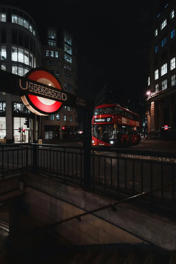 La imagen muestra una escena nocturna en una calle de Londres, destacando elementos icónicos de la ciudad. En el primer plano, se ve una entrada al metro (Underground) con el distintivo letrero circular rojo, blanco y azul. A la derecha del letrero, está el nombre de la estación: “Monument Station”. La entrada está rodeada por una barandilla negra metálica que delimita el acceso al transporte subterráneo.  En el fondo, se aprecia un clásico autobús rojo de dos pisos, característico de Londres, iluminado por las luces de la calle y de los edificios circundantes. Las edificaciones tienen grandes ventanas que reflejan la luz, añadiendo un contraste interesante entre el ambiente oscuro y las áreas iluminadas.  La escena transmite una atmósfera vibrante, típica de una ciudad activa incluso de noche, con un diseño urbano bien definido y elementos que refuerzan la identidad londinense.