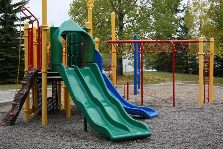 La imagen muestra un área de juegos infantiles al aire libre, equipada con una estructura de colores vivos diseñada para el entretenimiento de los niños. En el centro, destacan dos toboganes paralelos de color verde, con un diseño ancho y seguro. Junto a ellos, se encuentra un tobogán azul que añade variedad al conjunto.  La estructura principal está construida con postes de color amarillo y detalles en rojo, lo que le da un aspecto llamativo y alegre. Incluye otros elementos como un panel de juego interactivo en forma de “Tres en Raya” y una red negra que sirve como acceso o espacio de escalada. A la derecha, hay un conjunto de barras colgantes en color rojo, diseñadas para juegos de equilibrio y fuerza.  El suelo del parque está cubierto de grava, proporcionando un entorno seguro y resistente. Al fondo, se aprecian árboles verdes que enmarcan el espacio, creando un ambiente natural y agradable. Este parque parece un lugar ideal para que los niños disfruten y desarrollen sus habilidades físicas mientras se divierten.