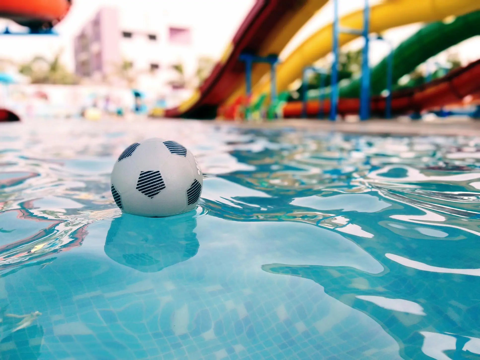La imagen muestra una pelota flotando en el agua de una piscina. La pelota, de diseño clásico con patrones hexagonales en blanco y negro que imitan el estilo de un balón de fútbol, está parcialmente sumergida, creando suaves ondulaciones en la superficie del agua.  El agua de la piscina es cristalina, con tonos azules y reflejos de luz que generan un efecto visual dinámico y relajante. Al fondo, desenfocado, se intuyen colores vibrantes, probablemente pertenecientes a elementos decorativos o accesorios cercanos a la piscina, que aportan un toque alegre a la escena.  La imagen transmite una sensación de diversión, ocio y calma, evocando un día relajado junto a la piscina. Ideal para representar actividades recreativas o momentos de descanso en un ambiente acuático.
