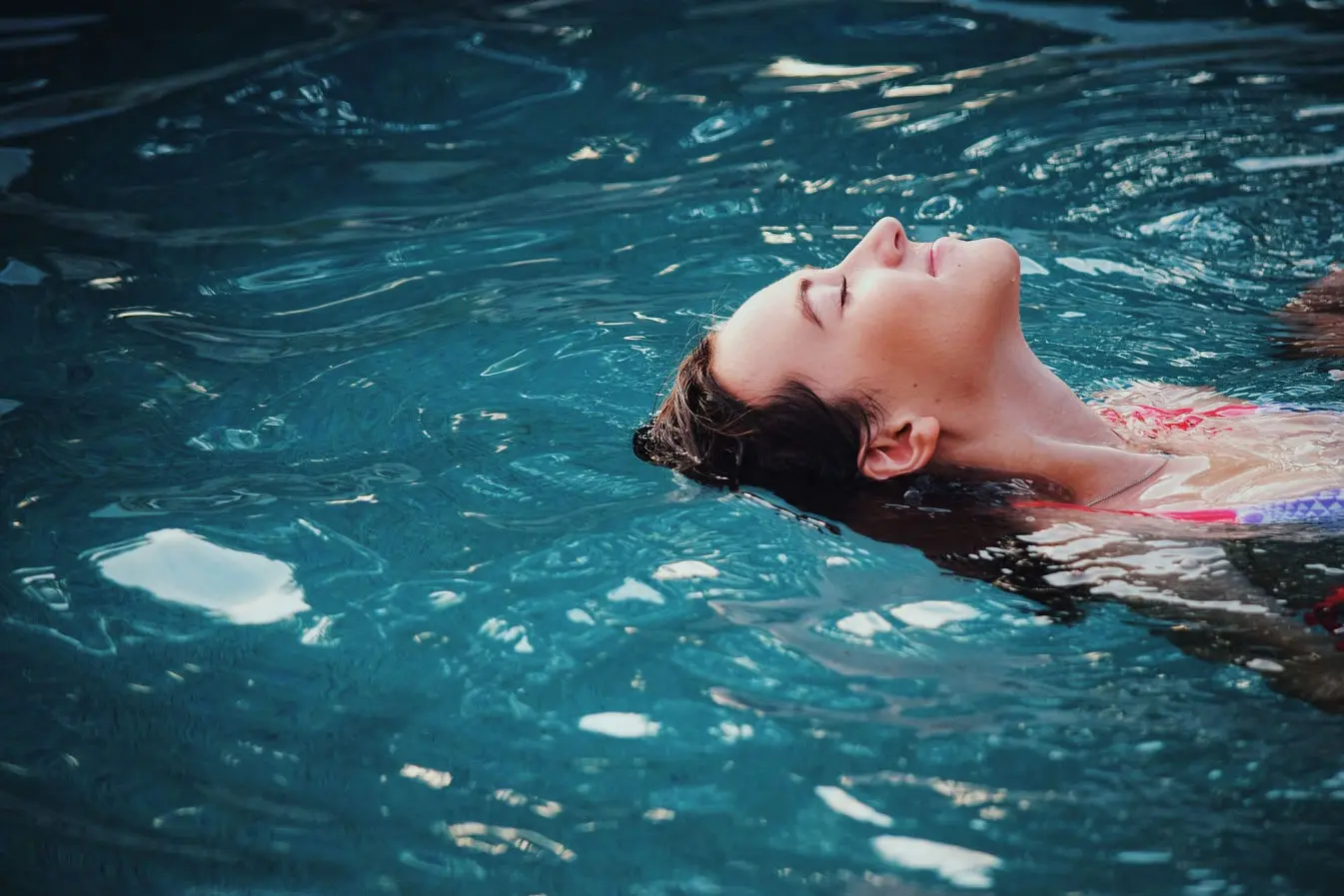 La imagen muestra a una mujer flotando relajadamente en el agua, con los ojos cerrados y una expresión de calma y serenidad en su rostro. Su cabeza está parcialmente sumergida, mientras su rostro y cuello emergen suavemente del agua, destacando por la luz natural que ilumina la escena. El agua, de un tono azul profundo, refleja sutiles destellos y ondulaciones que añaden dinamismo y textura a la composición.  La postura de la mujer, con la cabeza hacia atrás y el cuerpo relajado, transmite una sensación de paz y desconexión, probablemente disfrutando de un momento de descanso o meditación. La imagen evoca tranquilidad y armonía, siendo ideal para representar temas relacionados con el bienestar, la relajación o el contacto con la naturaleza.