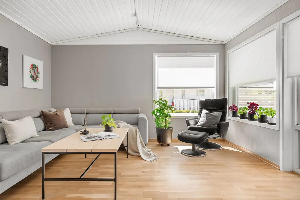 La imagen muestra un salón moderno y luminoso decorado con un estilo minimalista y acogedor. A la izquierda, hay un sofá en forma de “L” tapizado en color gris claro, decorado con cojines en tonos neutros y uno de color marrón metálico que aporta contraste. Frente al sofá, se encuentra una mesa de centro rectangular con una estructura de metal negro y una superficie blanca, sobre la cual descansan un libro, una planta y un candelabro sencillo, que refuerzan la sensación de simplicidad y orden.  A la derecha, junto a una gran ventana que permite la entrada de luz natural, hay un sillón reclinable negro con reposapiés a juego, acompañado de un cojín que lo hace aún más cómodo. En el alféizar de la ventana y en el suelo cercano, se disponen varias macetas con plantas verdes y flores, que añaden un toque de frescura y vida al espacio.  El suelo es de madera clara, y las paredes grises y blancas crean un ambiente equilibrado y sereno. El techo con paneles blancos y líneas rectas da una sensación de amplitud. En general, el espacio transmite una atmósfera de calma y comodidad, ideal para el descanso o la lectura.