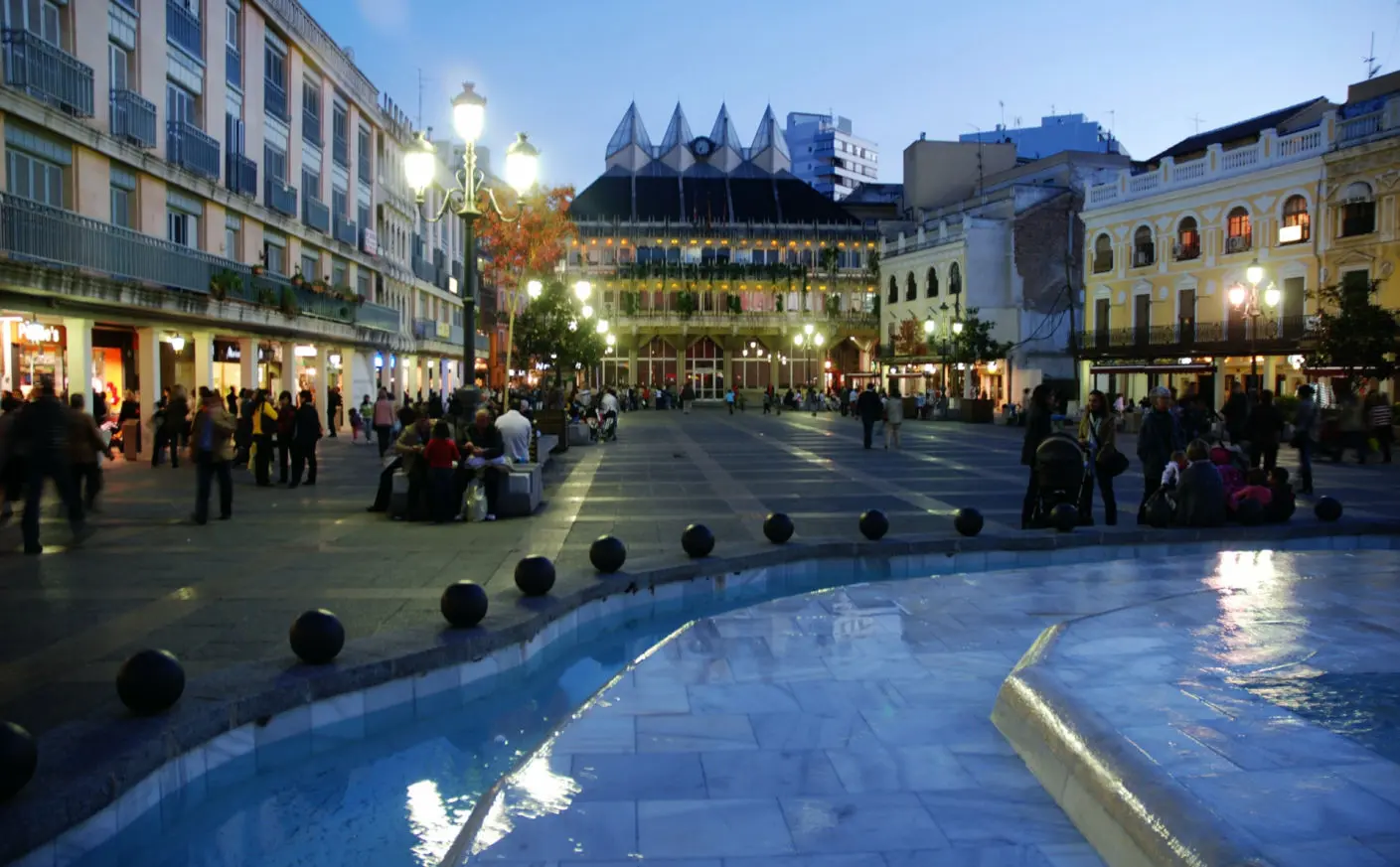 La imagen muestra una plaza pública en Ciudad Real durante la tarde-noche, iluminada por luces urbanas que crean una atmósfera animada y acogedora. En primer plano, se ve una fuente con agua que refleja las luces, añadiendo un toque de frescura y dinamismo al lugar.  A la izquierda, hay edificios de varios pisos con balcones y escaparates iluminados, lo que indica la presencia de tiendas y locales comerciales. La derecha está compuesta por construcciones más tradicionales, con fachadas decorativas y arcos que resaltan el encanto histórico de la zona.  En el centro de la plaza, varias personas pasean o se reúnen en grupos, lo que denota que es un espacio de encuentro y actividad social. Al fondo, se destaca un edificio moderno con cristales y luces que contrastan con la arquitectura clásica de los alrededores. La escena refleja la mezcla de tradición y modernidad que caracteriza a Ciudad Real, con un ambiente vibrante y acogedor.