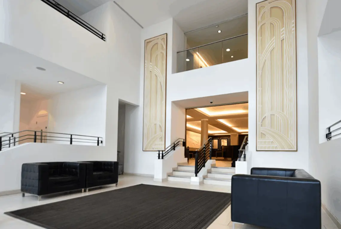 La imagen muestra un interior amplio y moderno, con un diseño minimalista y detalles que evocan el estilo art déco. Las paredes son predominantemente blancas, creando una atmósfera luminosa y elegante. En el centro, se observa una escalera que lleva a un nivel superior y una pasarela de vidrio que conecta las áreas superiores, añadiendo un toque contemporáneo.  A ambos lados de la escalera principal, hay barandillas negras que contrastan con el blanco de las paredes. A la derecha e izquierda se encuentran paneles decorativos de tonos beige y crema con patrones geométricos curvados, característicos del art déco. En el área de espera o recepción, se aprecian sofás de diseño moderno en color negro, colocados sobre una alfombra oscura que define el espacio.  La iluminación es cálida, y se aprecia tanto en el techo como en las áreas de paso, reforzando la sensación de sofisticación y funcionalidad. El diseño general combina elementos clásicos y modernos, creando un ambiente acogedor y profesional.