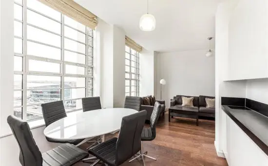 La imagen muestra un espacio interior luminoso y moderno, que combina un área de comedor y una sala de estar. A la izquierda, destaca una pared con amplios ventanales de estilo industrial, que permiten la entrada de abundante luz natural y ofrecen vistas de un paisaje urbano. Los ventanales están enmarcados por molduras blancas que complementan la estética limpia del espacio.  En el área de comedor, hay una mesa ovalada blanca rodeada por sillas de diseño moderno en color negro con respaldo acolchado. Este conjunto crea un contraste elegante con el suelo de madera cálida que se extiende por todo el espacio.  Al fondo, la sala de estar cuenta con un sofá gris oscuro decorado con cojines claros, que aportan comodidad y equilibrio al diseño. Una lámpara de pie blanca y una mesa de centro negra completan el mobiliario de la sala. A la derecha, se observa una encimera de cocina en color negro, que delimita visualmente el espacio, manteniendo la continuidad del diseño abierto.  El ambiente general es funcional y sofisticado, ideal para disfrutar de luz natural y comodidad en un entorno urbano.
