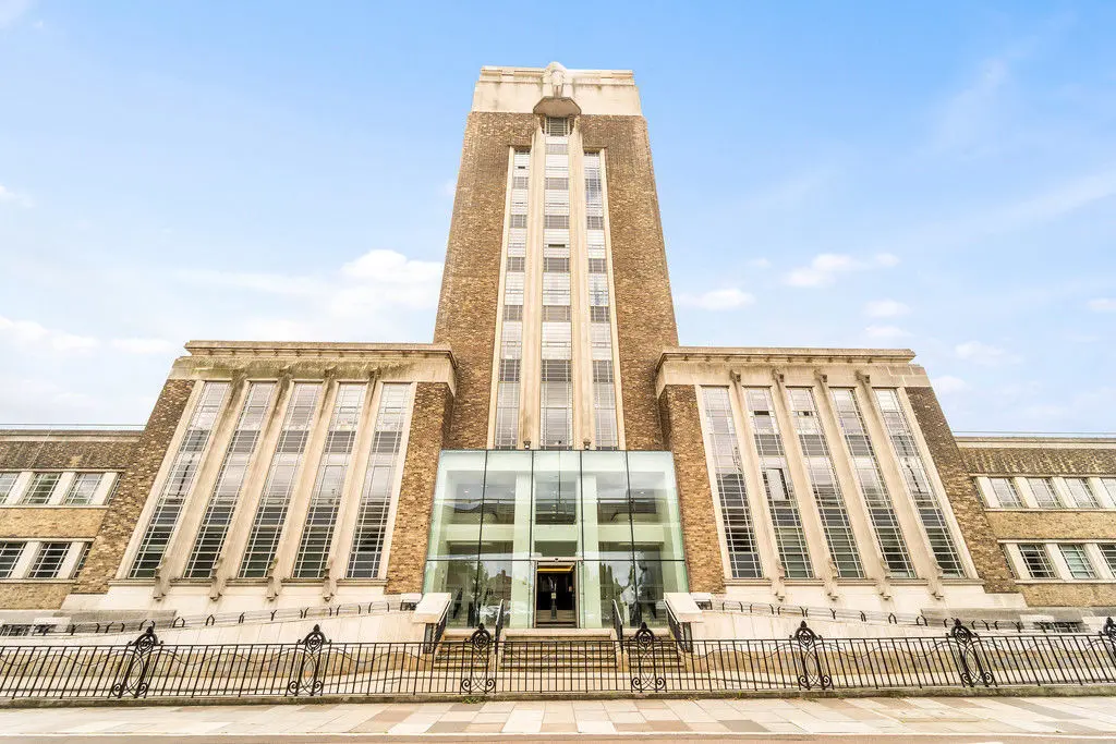 La imagen muestra un edificio de estilo arquitectónico moderno y simétrico, con elementos de líneas rectas y diseño geométrico. La estructura principal tiene una fachada de ladrillo con grandes ventanas verticales, enmarcadas por columnas y molduras de piedra clara, que le dan un aspecto monumental. En el centro, se encuentra una entrada con un diseño contemporáneo de vidrio y metal, que contrasta con el estilo clásico del resto del edificio. Este acceso está flanqueado por rampas y escaleras que conducen al nivel principal, con barandillas decorativas de hierro forjado. El cielo despejado resalta los detalles arquitectónicos del edificio. 